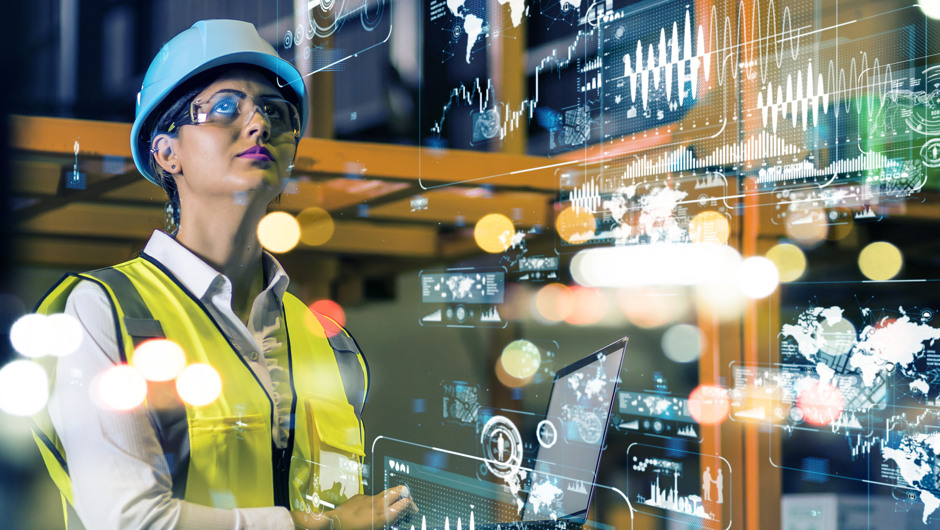 Kvinne i industrielt miljø med vernehjelm og refleksvest ser på digitale data og grafer som svever foran henne. Bildet illustrerer smart produksjon og datadrevet industri.