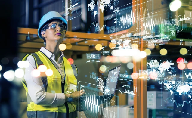 A woman in an industrial environment wearing a safety helmet and reflective vest is looking at digital data and graphs floating in front of her. The image illustrates smart manufacturing and data-driven industry.