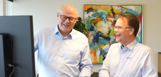 Two men standing in front of a computer monitor.