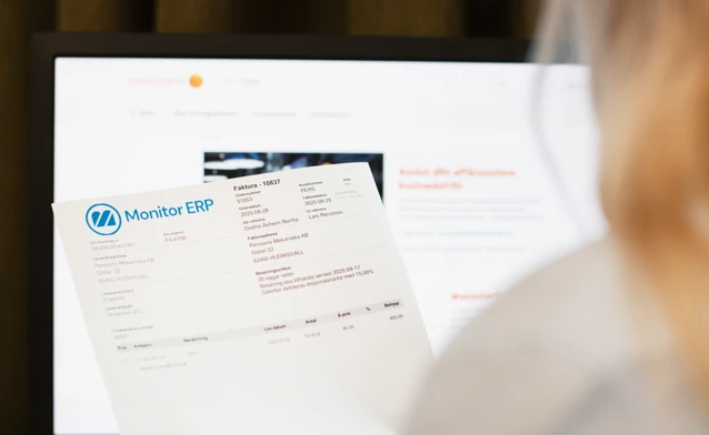 A woman holding an invoice from Monitor ERP in front of a computer screen.