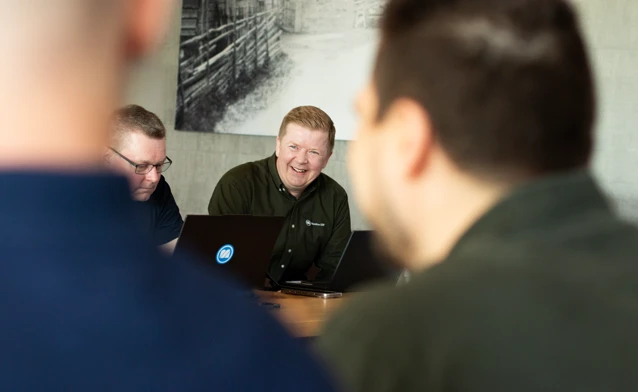 A group of men sitting around a laptop computer with a Monitor ERP logo. The men are happy and are smiling.