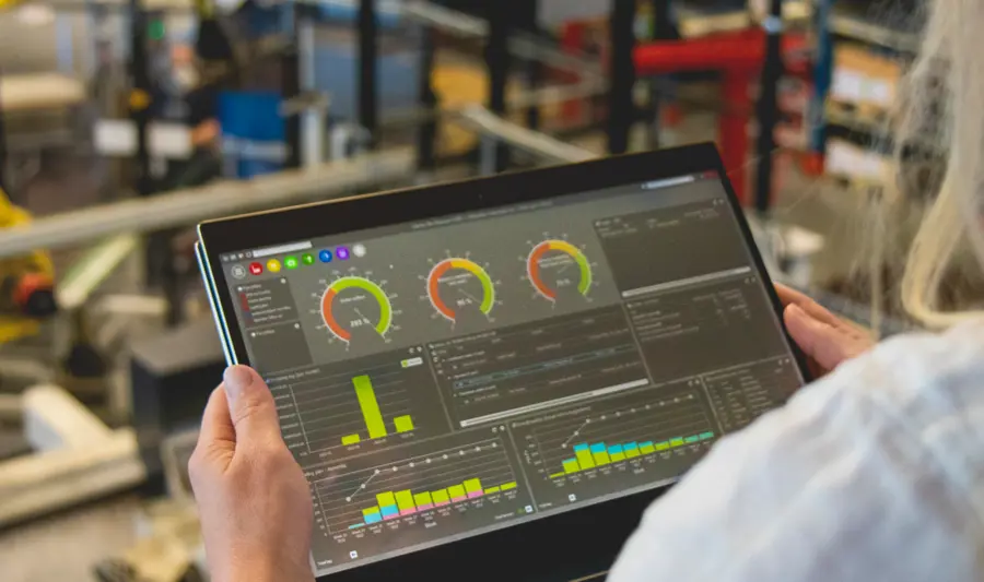 Kobieta trzymająca tablet z włączonym systemem Monitor ERP, w tle maszyny i roboty na hali produkcyjnej.
