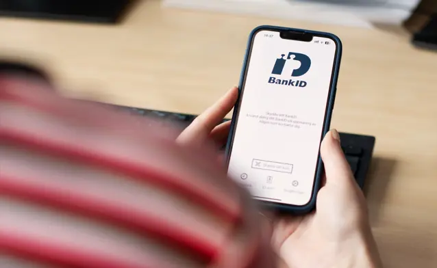 A person holding a cell phone over a desk with BankID visible on the screen.