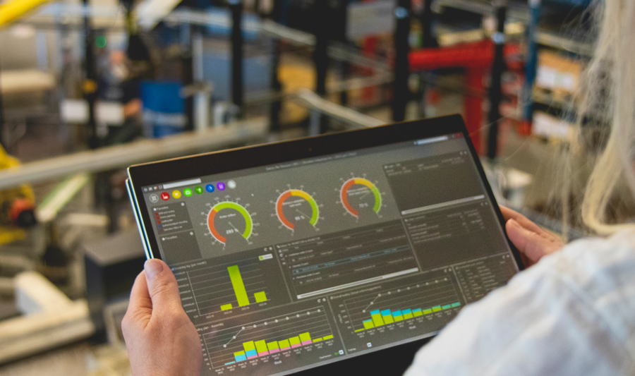 En person håller i en surfplatta i en fabriksmiljö där Monitor ERP-dashboard visas.