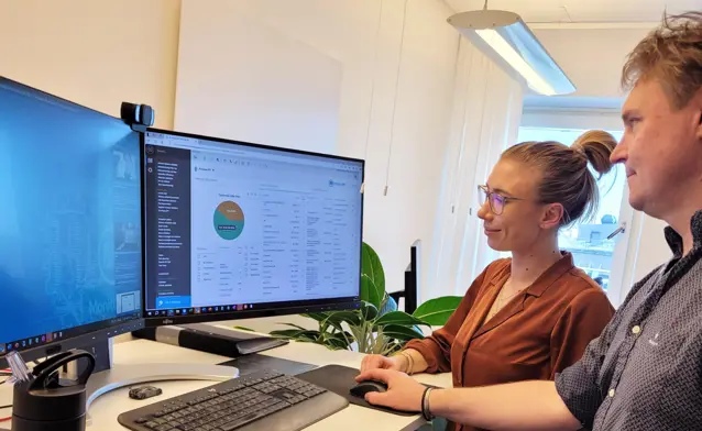 Two people are standing in front of a computer, where one is demonstrating and explaining how the Monitor ERP system works to the other.