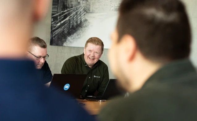 A group of men sitting around a laptop computer with a Monitor ERP logo. The men are happy and are smiling.