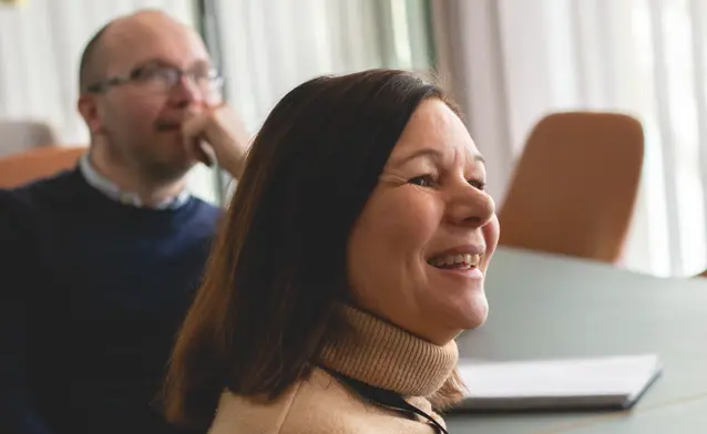 Två personer sitter vid ett bord i ett konferensrum och lyssnar på en presentation. 