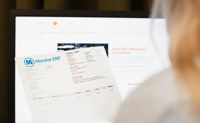 A woman holding an invoice from Monitor ERP in front of a computer screen.