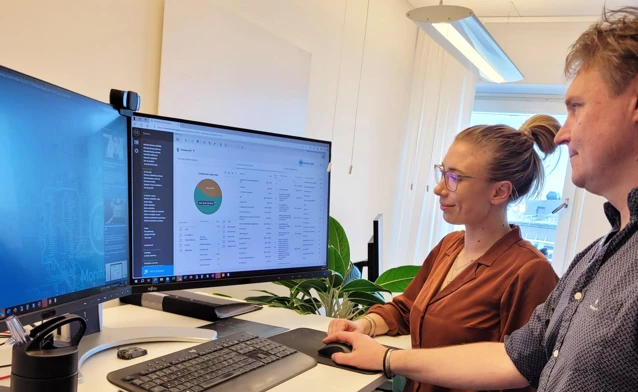 Two people are standing in front of a computer, where one is demonstrating and explaining how the Monitor ERP system works to the other.