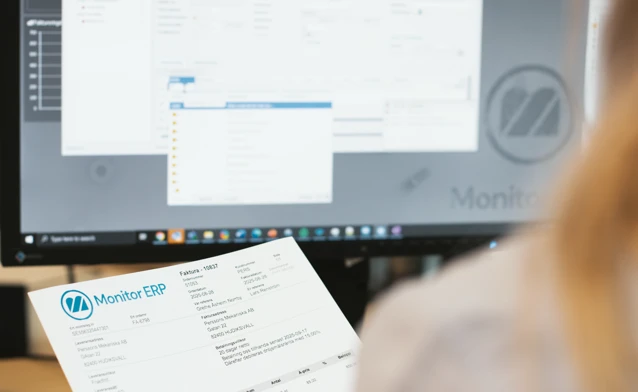 A person holding an invoice in front of a computer screen while checking information in Monitor ERP.