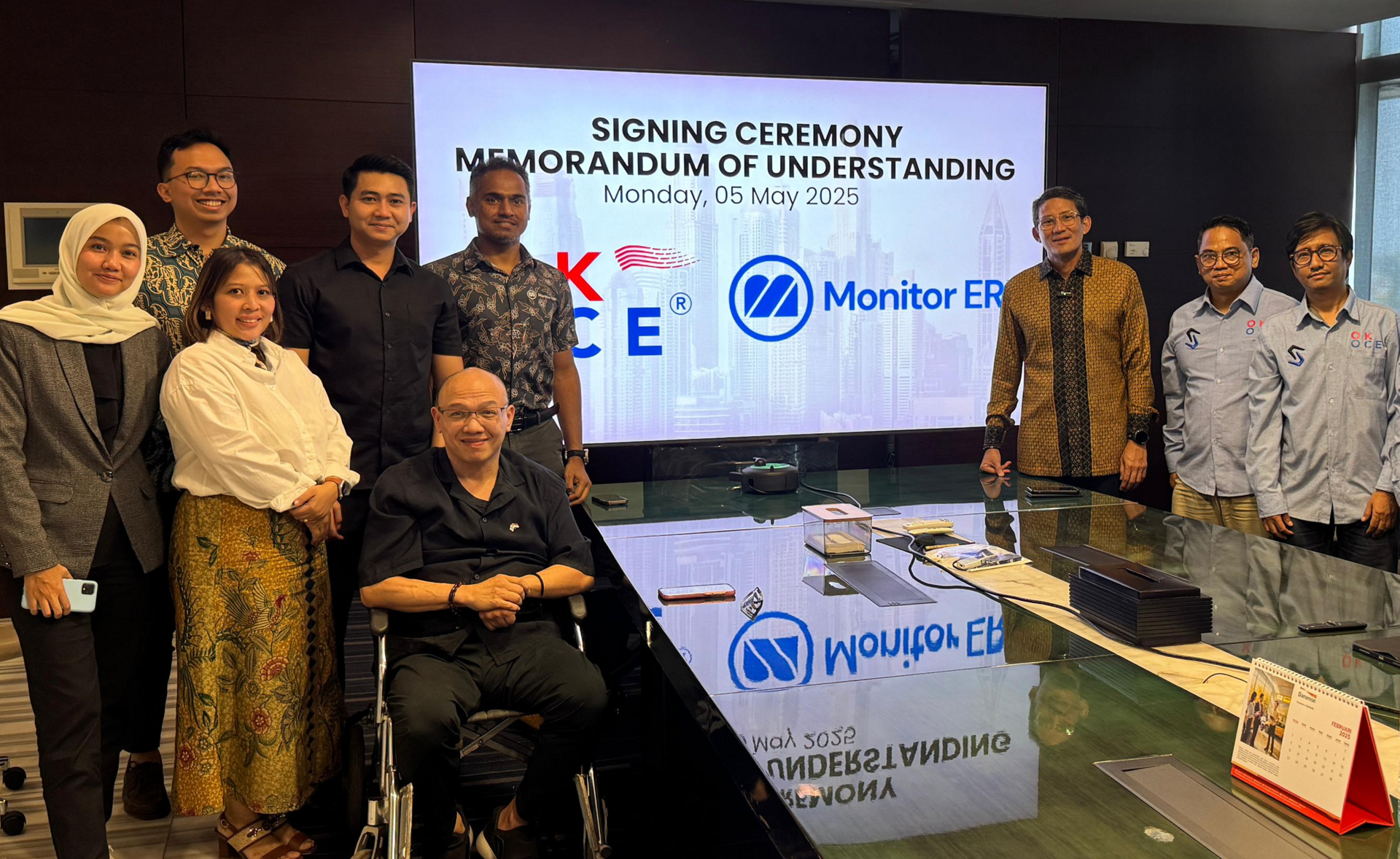 Group photo of professionals from OK OCE and Monitor ERP Indonesia, standing and seated around a conference table in a modern meeting room, with a large screen displaying 'Signing Ceremony Memorandum of Understanding'