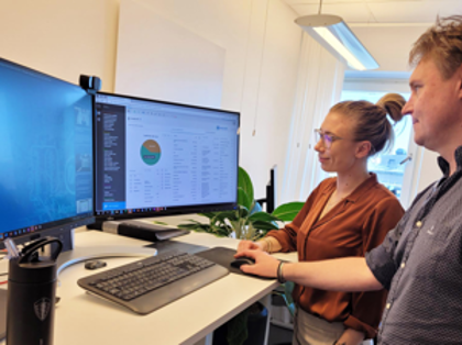 Two people are standing in front of a computer, where one is demonstrating and explaining how the Monitor ERP system works to the other.