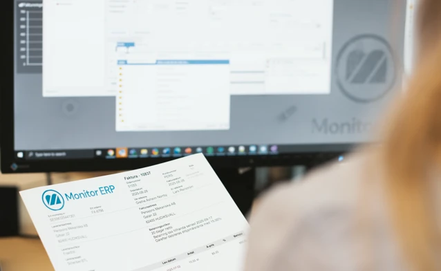 A person holding an invoice  in front of a computer screen while checking information in Monitor ERP.