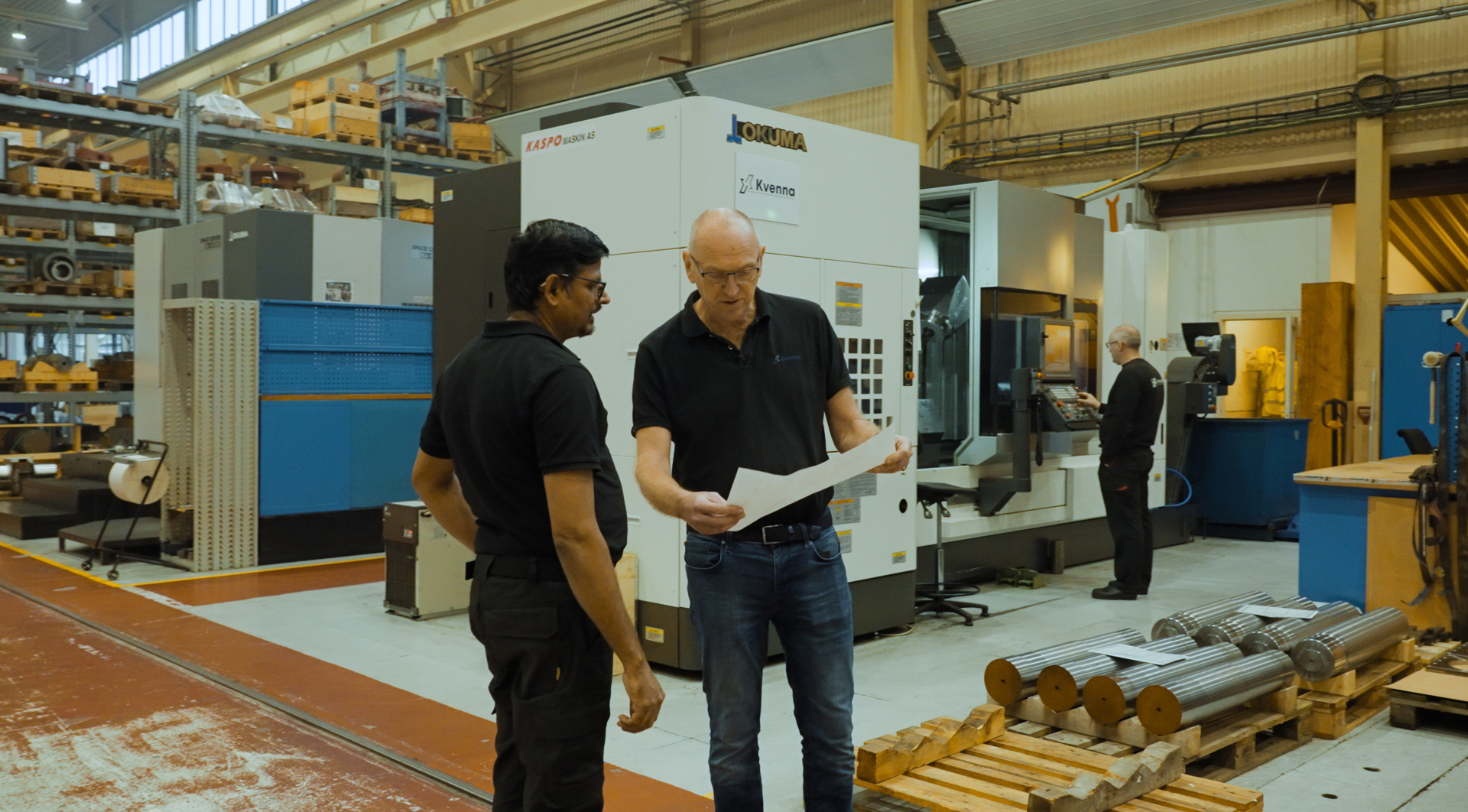 To industriarbeidere diskuterer en tegning foran en stor CNC-maskin i en produksjonshall. Hyller med metallkomponenter og flere arbeidsstasjoner er synlige i bakgrunnen, hvor en annen operatør arbeider ved en maskin.