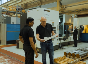 To industriarbeidere diskuterer en tegning foran en stor CNC-maskin i en produksjonshall. Hyller med metallkomponenter og flere arbeidsstasjoner er synlige i bakgrunnen, hvor en annen operatør arbeider ved en maskin.