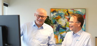 Two men standing in front of a computer monitor.